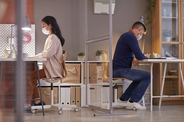 Vista laterale verticale di due persone che indossano maschere in ufficio mentre si lavora alle scrivanie in cubicoli separati post pandemia, spazio di copia
