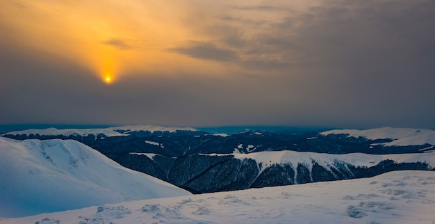 Vista mistica delle colline innevate in una valle di montagna in inverno contro il sole dorato al tramonto con un cielo nebbioso