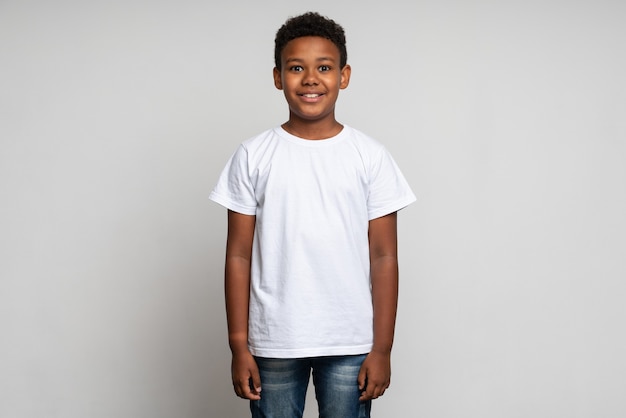 Vista orizzontale di un ragazzino carino con un'elegante pettinatura riccia in maglietta bianca in piedi e che guarda l'obbiettivo con faccia felice. Colpo dello studio dell'interno isolato su fondo bianco