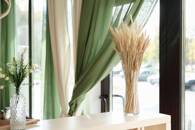 vista parziale degli interni eleganti del bar o della terrazza con decorazioni naturali spiga di grano in un vaso di vetro su un tavolo di legno chiaro tende verdi e fiori finestra con vista su una strada