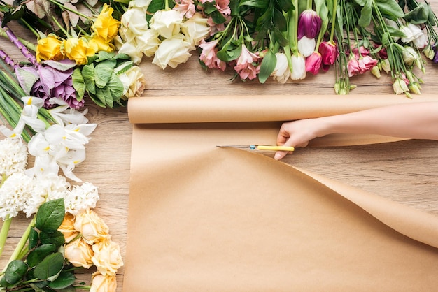 Vista parziale del fiorista che taglia la carta artigianale con le forbici vicino ai fiori su una superficie di legno