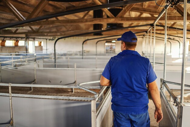 Vista posteriore del contadino in abiti da lavoro che cammina intorno al fienile vuoto e controlla la pulizia