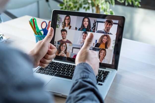 Vista posteriore del dipendente di sesso maschile che fa un'approvazione gestuale con le dita del pollice mentre parla in videochiamata con diversi colleghi durante il briefing online con il laptop a casa.