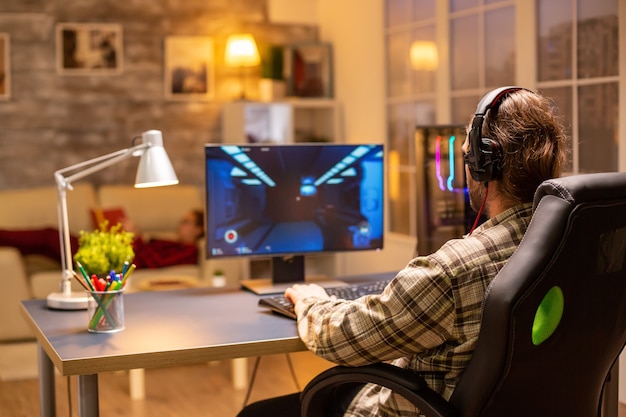 Vista posteriore del giocatore maschio che gioca a uno sparatutto sul suo potente computer PC a tarda notte nel soggiorno.