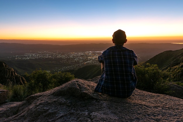 Vista posteriore del giovane seduto su una scogliera a guardare il tramonto dietro le luci della città