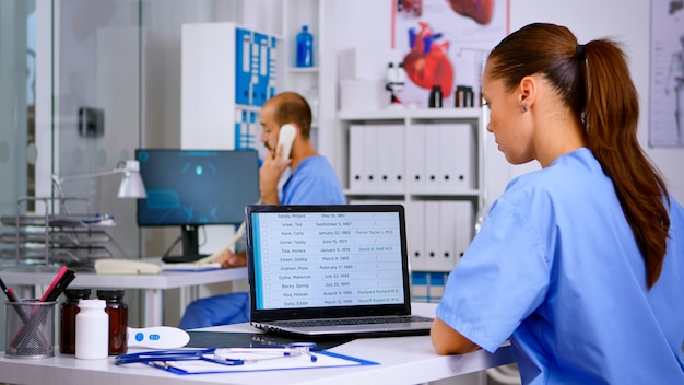 Vista posteriore del medico residente che controlla gli appuntamenti sul laptop mentre il collega risponde al telefono offrendo consulenza medica al paziente remoto, assistente che lavora nella clinica ospedaliera