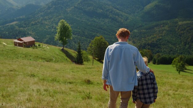 Vista posteriore del padre dai capelli rossi con il figlio che cammina sulla collina verde giornata di sole Giovane genitore spensierato che abbraccia il ragazzo biondo che scende dal pendio della montagna Uomo sconosciuto che trascorre le vacanze estive con il bambino sulla natura