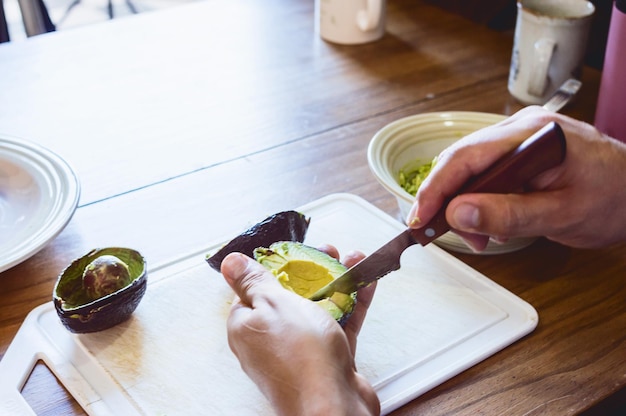 Vista posteriore del primo piano delle mani maschili che tagliano le fette di avocado con un coltello su un tagliere bianco