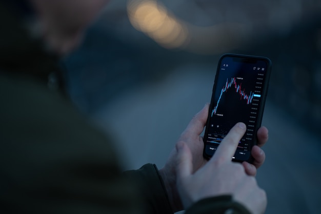 Vista posteriore del trader che monitora i dati del mercato azionario nell'app mobile per il trading online e gli investimenti su smartphone stando in piedi all'aperto, messa a fuoco selettiva sullo schermo tattile a mano con grafico grafico forex