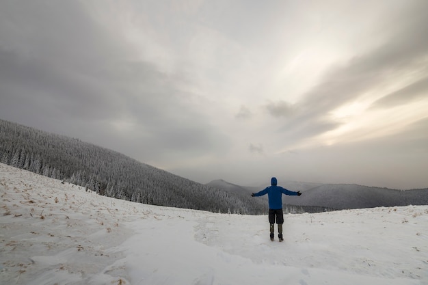 Vista posteriore dell'escursionista turistico in abiti caldi con lo zaino in piedi con le braccia alzate sulla radura coperta di neve sulla montagna della foresta di abete rosso e sullo sfondo dello spazio della copia del cielo nuvoloso.