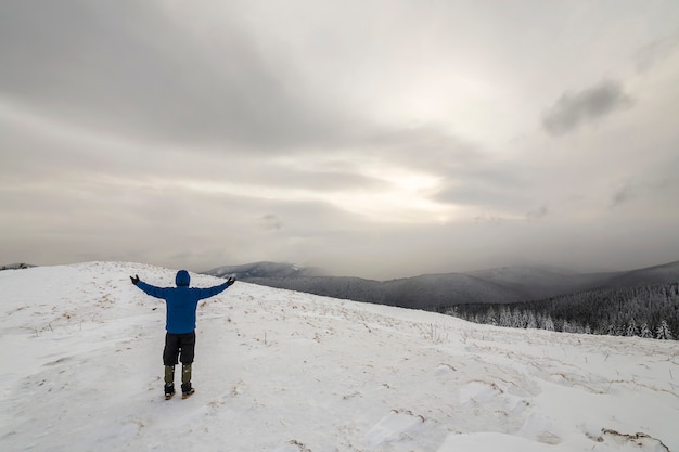 Vista posteriore dell'escursionista turistico in abiti caldi con zaino in piedi con le braccia alzate sulla radura ricoperta di neve sulla montagna della foresta di abete rosso e sfondo spazio copia cielo nuvoloso