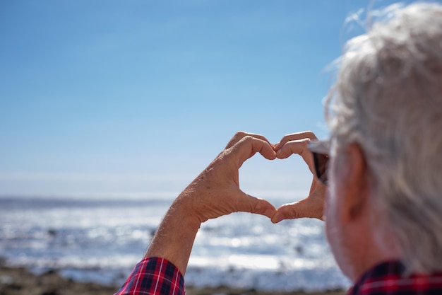 Vista posteriore dell'uomo anziano in camicia a scacchi in piedi sulla spiaggia che fa la forma del cuore con le mani