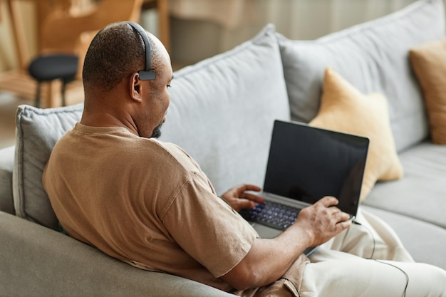 Vista posteriore dell'uomo di colore adulto che utilizza il laptop con l'auricolare mentre si lavora in una riunione online dal poliziotto di casa