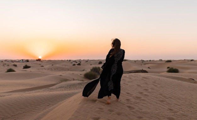 Vista posteriore della bella donna misteriosa in abito lungo nero arabo tradizionale si trova nel deserto al tramonto