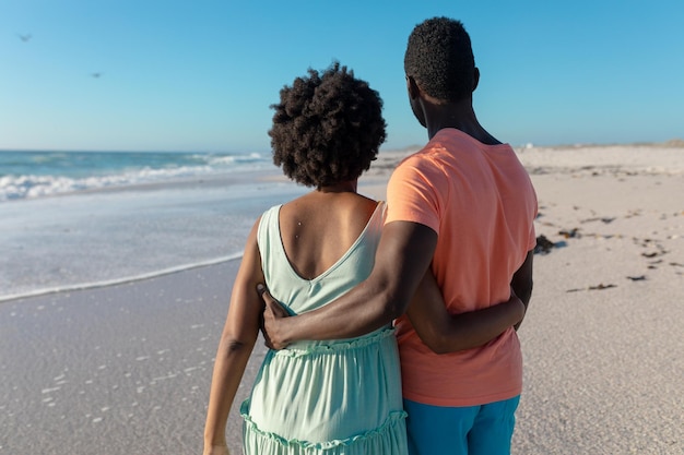 Vista posteriore della coppia afroamericana in piedi con le braccia intorno alla spiaggia durante le vacanze estive