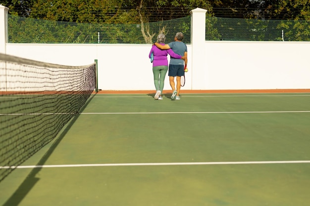 Vista posteriore della coppia anziana birazziale con le braccia intorno a camminare sul campo da tennis durante la giornata di sole