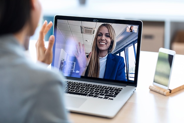 Vista posteriore della dipendente che saluta e parla in videochiamata con il suo collega durante il briefing online con il laptop a casa.