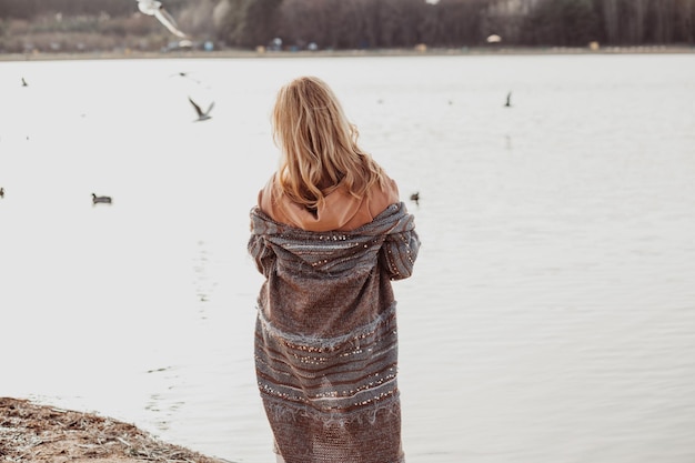 Vista posteriore della donna con lunghi capelli biondi ondulati che indossa cardigan lungo felpa marrone in piedi sulla spiaggia sabbiosa del fiume