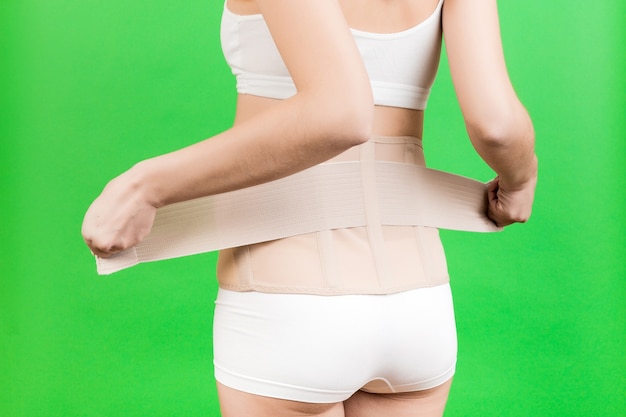 Vista posteriore della donna incinta in biancheria intima che veste corsetto ortopedico per far andare via il mal di schiena a sfondo verde con spazio di copia. Primo piano del concetto di cintura di supporto addominale ortopedico.