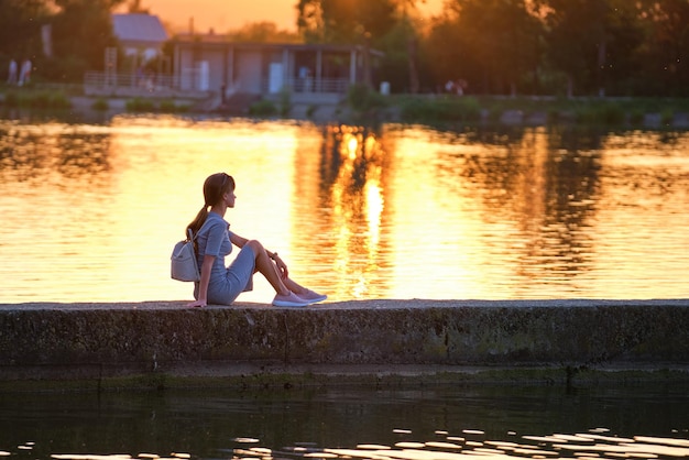 Vista posteriore della donna sola seduta sulla riva del lago in una calda serata. Concetto di solitudine e relax.