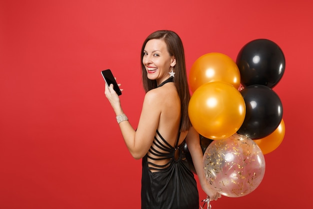 Vista posteriore della donna sorridente che guarda indietro tenendo, utilizzando il telefono cellulare con schermo vuoto nero vuoto, mongolfiere isolate su sfondo rosso. Felice anno nuovo, concetto di festa di compleanno mockup.