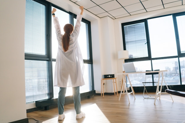 Vista posteriore della dottoressa in camice bianco che allunga le mani sullo sfondo della finestra in una giornata di sole.