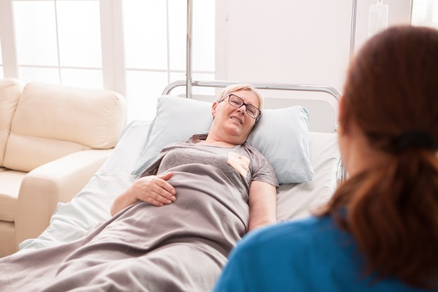 Vista posteriore della dottoressa in casa di cura che parla con una donna anziana sdraiata a letto.