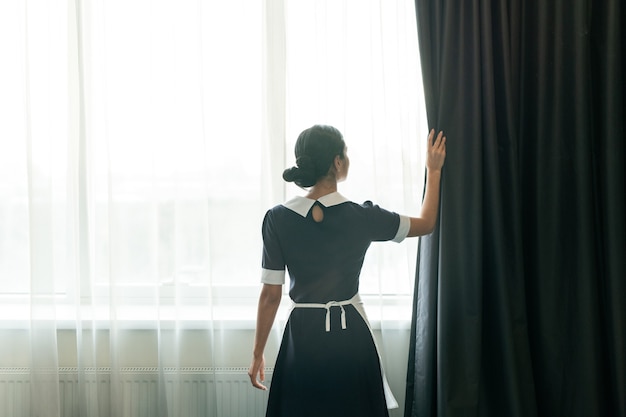 Vista posteriore della giovane cameriera in piedi vicino alla grande finestra