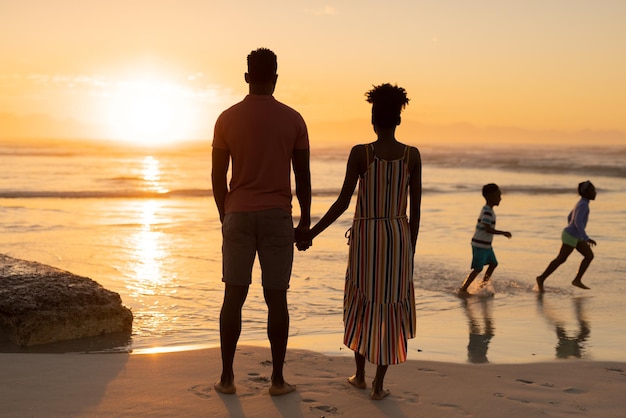 Vista posteriore della giovane coppia afroamericana che guarda i bambini giocosi che corrono sulla spiaggia contro il cielo