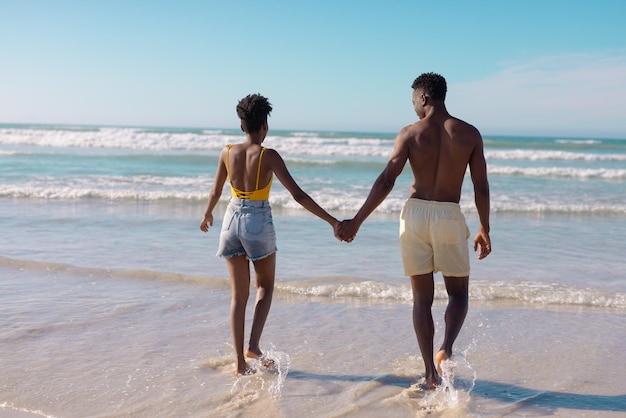 Vista posteriore della giovane coppia afroamericana che si tiene per mano e cammina verso il mare contro il cielo