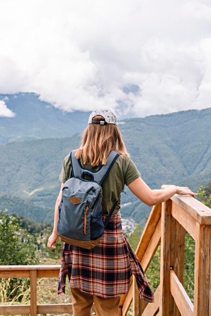 Vista posteriore della giovane donna bionda in berretto con zaino e camicia a quadri guardando la vista delle montagne