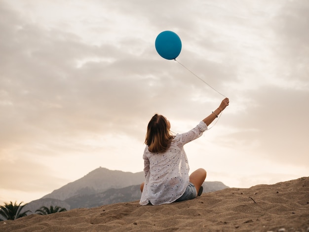 Vista posteriore della giovane donna seduta sulla spiaggia con palloncino di elio blu.