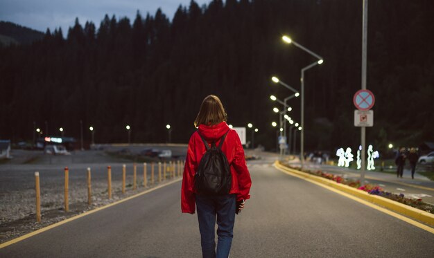 Vista posteriore della giovane ragazza turistica sulla strada che va alla foresta di montagna nelle luci della strada