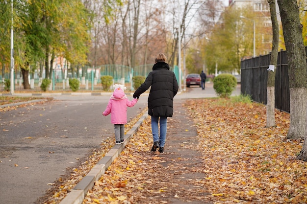 Vista posteriore della madre e della sua piccola figlia che camminano lungo la strada della città Donna che conduce il bambino per maniglia lungo il marciapiede nella stagione autunnale