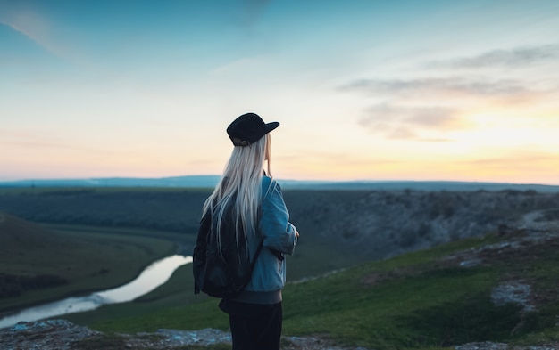 Vista posteriore della ragazza bionda con zaino nero e cappuccio. Giovane viaggiatore guardando il tramonto dalle colline.