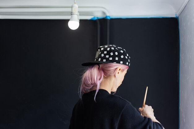 Vista posteriore della ragazza con i capelli rosa, pittura murale con pennello e colore bianco, aggiornamento dell'appartamento. Indossare berretto nero e maglione.