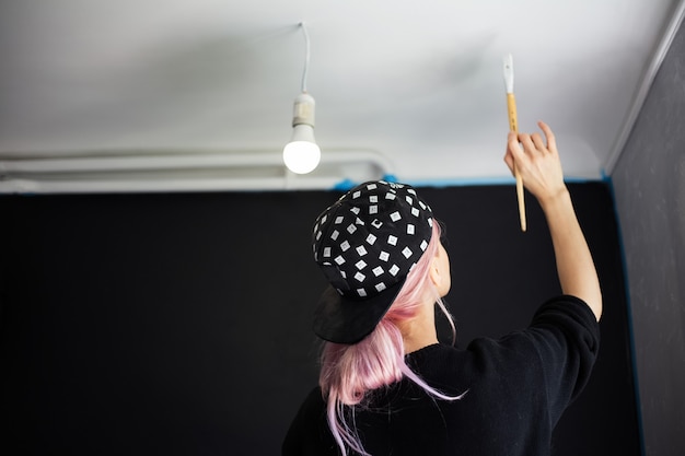 Vista posteriore della ragazza con i capelli rosa, pittura murale con pennello e colore bianco, aggiornamento dell'appartamento. Indossare berretto nero e maglione.