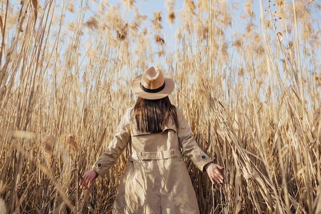 Vista posteriore della ragazza in trench e cappello in erba alta pampa con cielo blu sullo sfondo