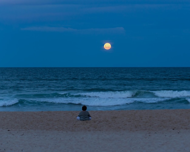 Vista posteriore della vecchia donna seduta sulla spiaggia a guardare la luna piena