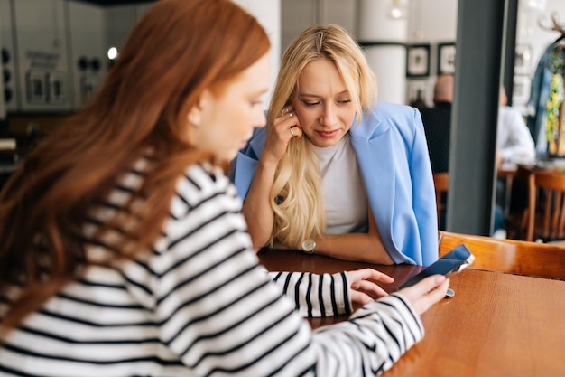 Vista posteriore di due belle giovani donne sedute a tavolo con il caffè al caffè vicino alla finestra che conversano e si mostrano qualcosa sui telefoni cellulari