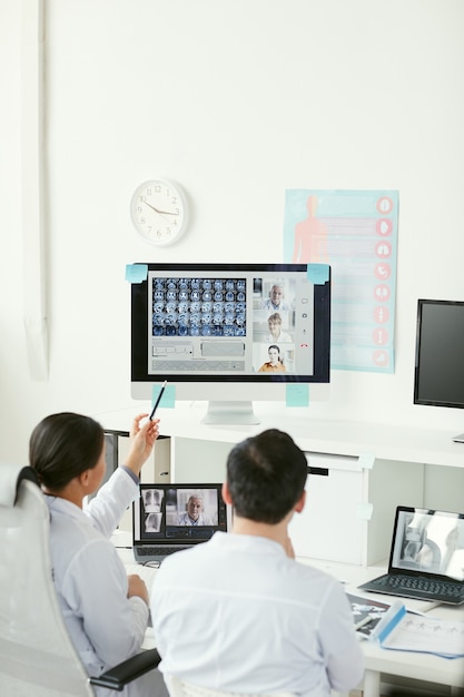 Vista posteriore di due medici seduti al tavolo ed esaminando le immagini radiografiche sul computer insieme ai loro colleghi