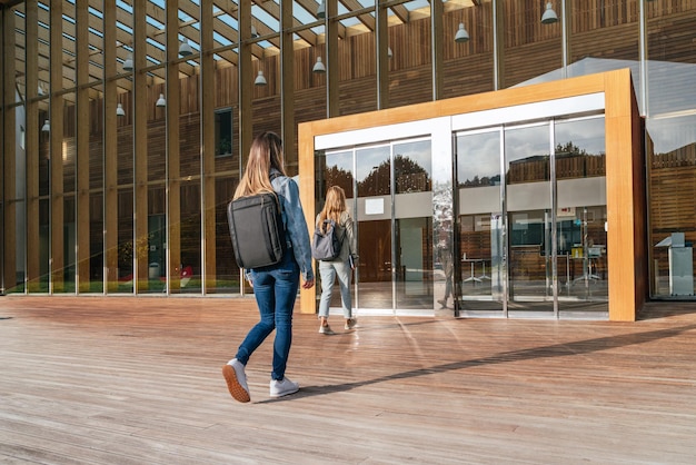 Vista posteriore di due studenti universitari irriconoscibili che entrano nell'università per frequentare le lezioni in una giornata di sole.