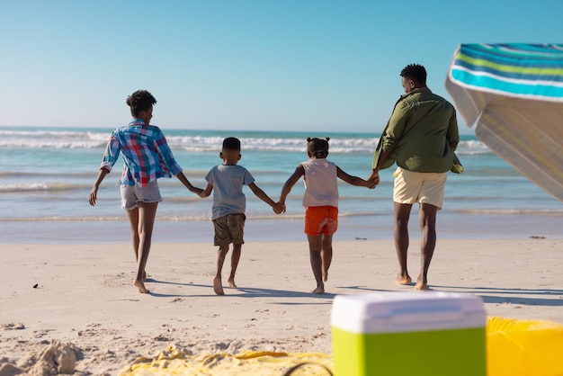 Vista posteriore di giovani genitori afroamericani che tengono le mani dei bambini che corrono in spiaggia contro il cielo