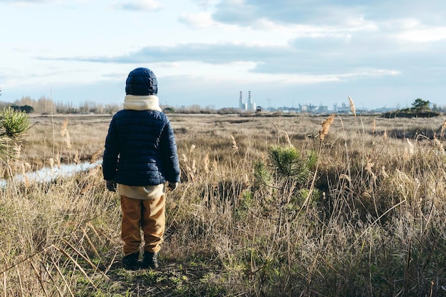 Vista posteriore di un bambino che indossa abiti invernali in piedi di fronte al paesaggio selvaggio e alle fabbriche sullo sfondo Concetto di attivismo e protezione ambientale Futuro dell'infanzia per le nuove generazioni