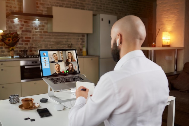 Vista posteriore di un dipendente di sesso maschile con gli auricolari che sta prendendo appunti su una videoconferenza aziendale su un laptop. Un uomo calvo con la barba durante un incontro online con i suoi colleghi a casa