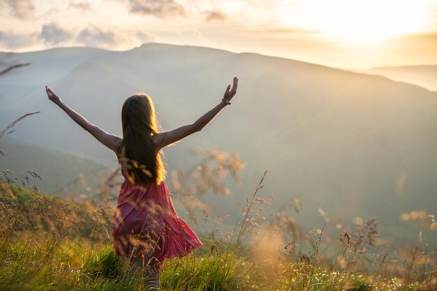 Vista posteriore di un'escursionista donna felice in abito rosso in piedi sulla collina erbosa in una serata ventosa in montagne autunnali con le braccia tese godendo della vista della natura al tramonto.