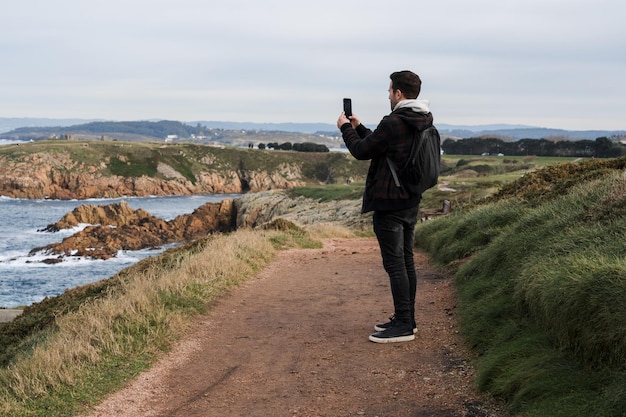 Vista posteriore di un giovane che fotografa il paesaggio con il suo cellulare in piedi su una scogliera