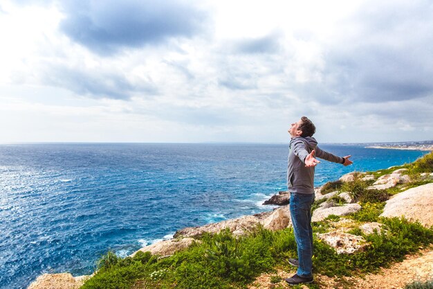 Vista posteriore di un giovane ragazzo in piedi su un bordo di una scogliera rocciosa al mare con le braccia aperte contro un paesaggio marino