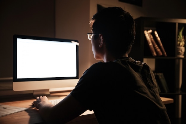 Vista posteriore di un giovane serio con gli occhiali seduto e che lavora con un computer a schermo vuoto a casa durante la notte