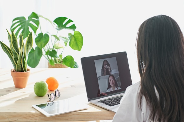 Vista posteriore di un medico asiatico che conduce una consultazione online con un paziente sul portatile mentre è seduto a tavola con un tablet in clinica